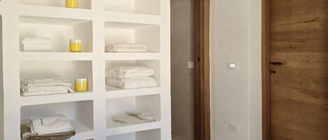 Une chambre avec un lit, une porte en bois et une étagère intégrée où sont rangées des serviettes pliées et des bougies.