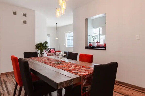 Un espace repas comprenant une table en bois, des chaises rouges et noires, un chemin de table à motifs et un luminaire suspendu.