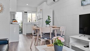Una cocina moderna con armarios blancos, un comedor con sillas de madera y una mesa, y un televisor sobre un soporte blanco.