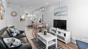 Una sala de estar moderna con un sofá de cuero negro, una mesa de centro blanca y un televisor de pantalla plana sobre un soporte blanco. Hay una zona de comedor con una mesa y sillas de madera, y una cocina al fondo.