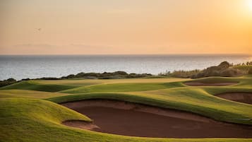 A golf course with sand bunkers and a view of the ocean at sunset.