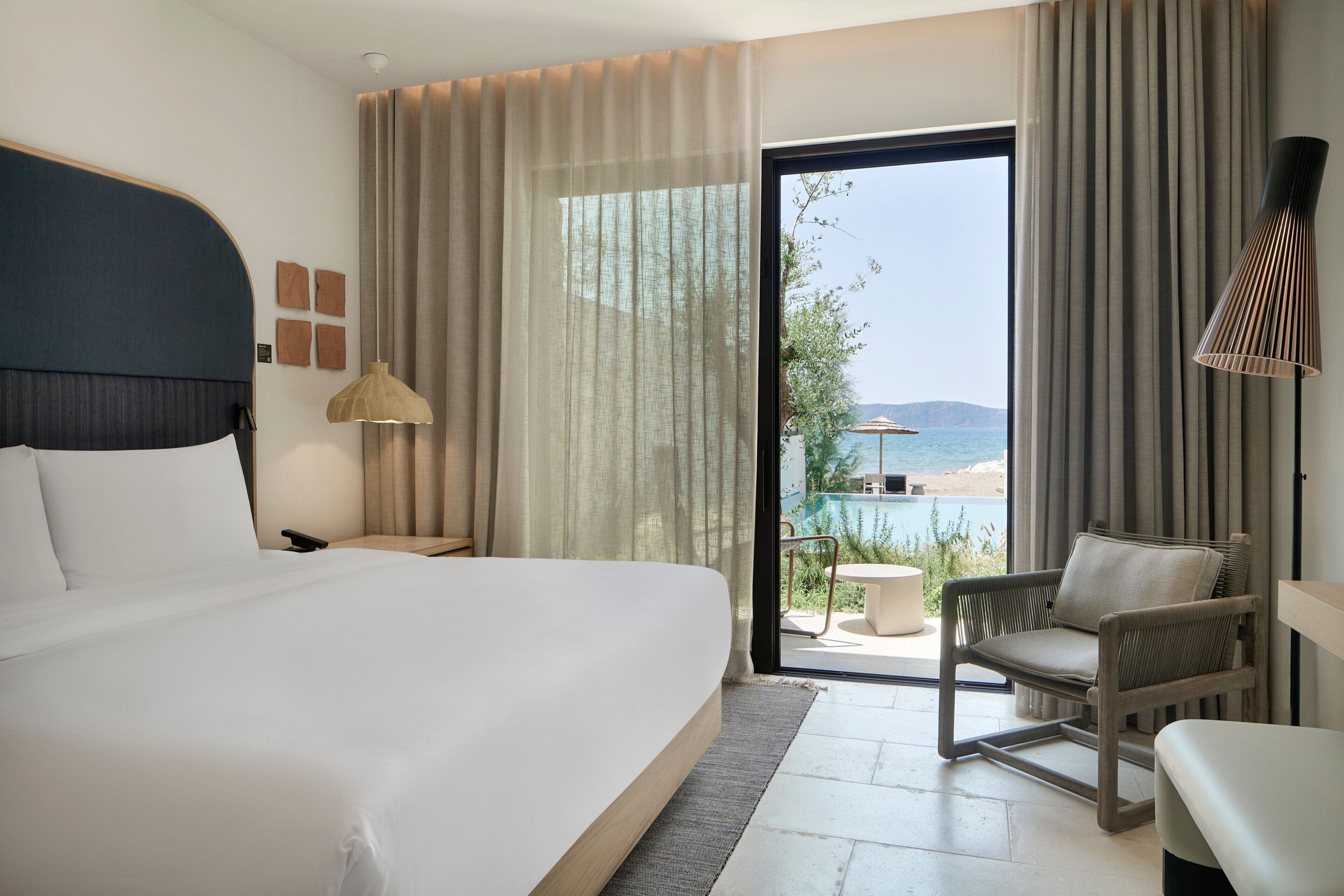 A hotel room with a large bed, a chair, a small table, and a view of the beach through the window.