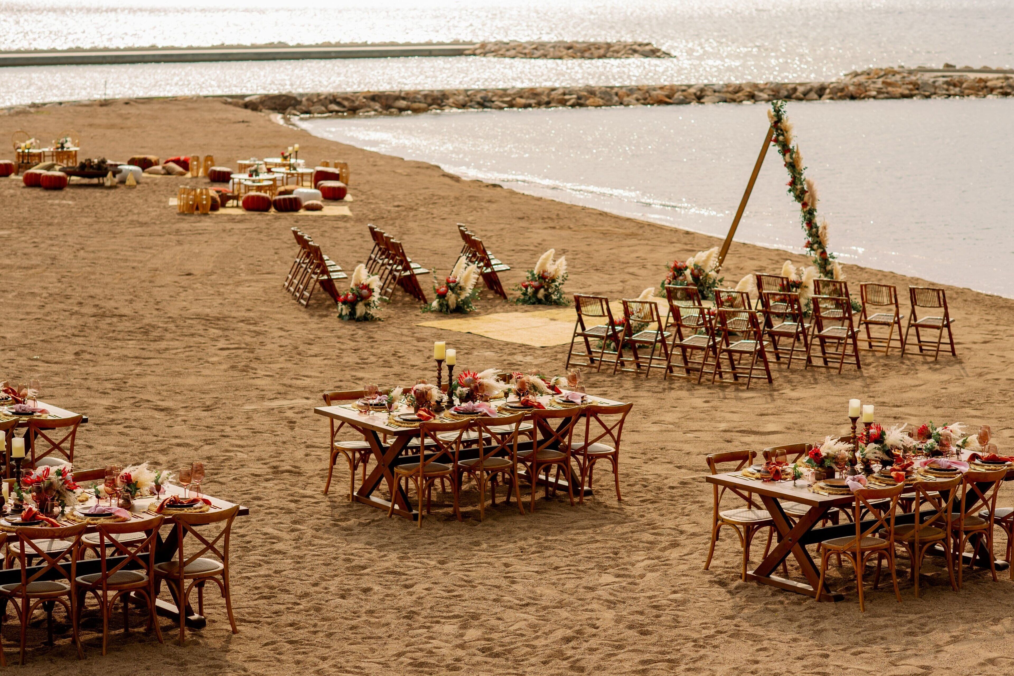 A beach wedding setup with tables and chairs arranged for guests, a decorated aisle, and a backdrop of the ocean.