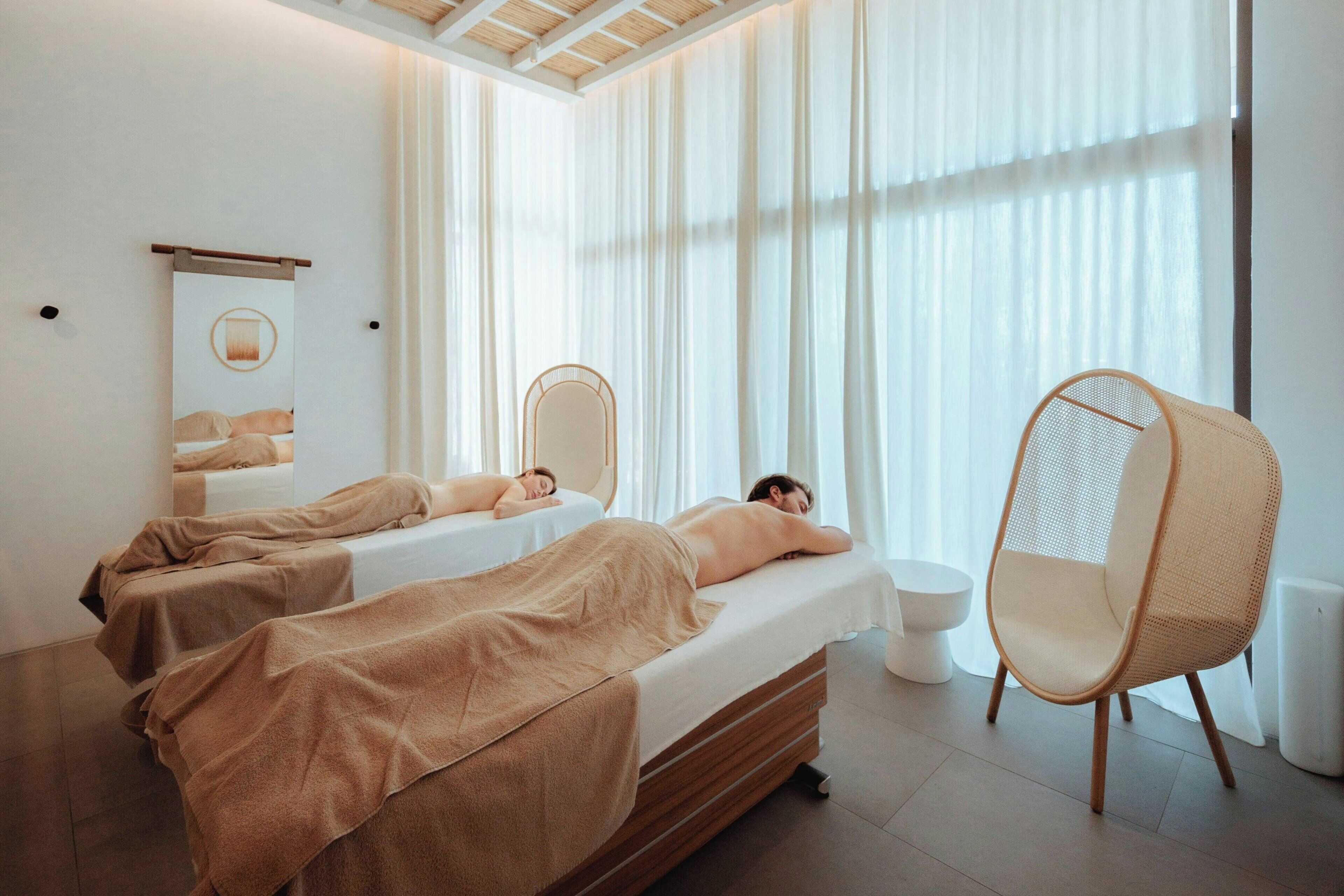 A spa room with two beds, a large window with sheer curtains, a wooden ceiling, and a rattan chair.