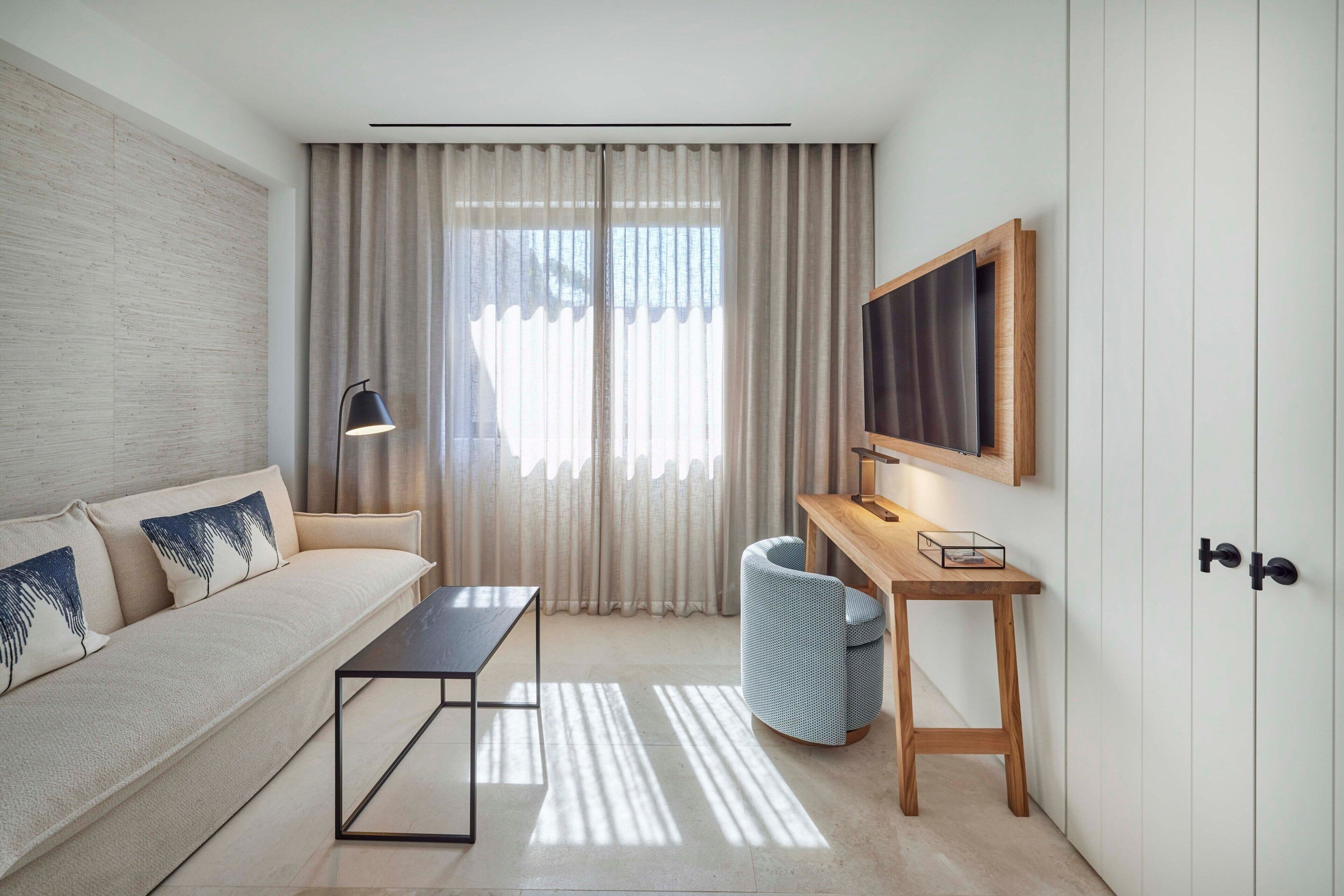 A modern, well-lit room with a sofa, a desk, a lamp, and a large window with curtains.