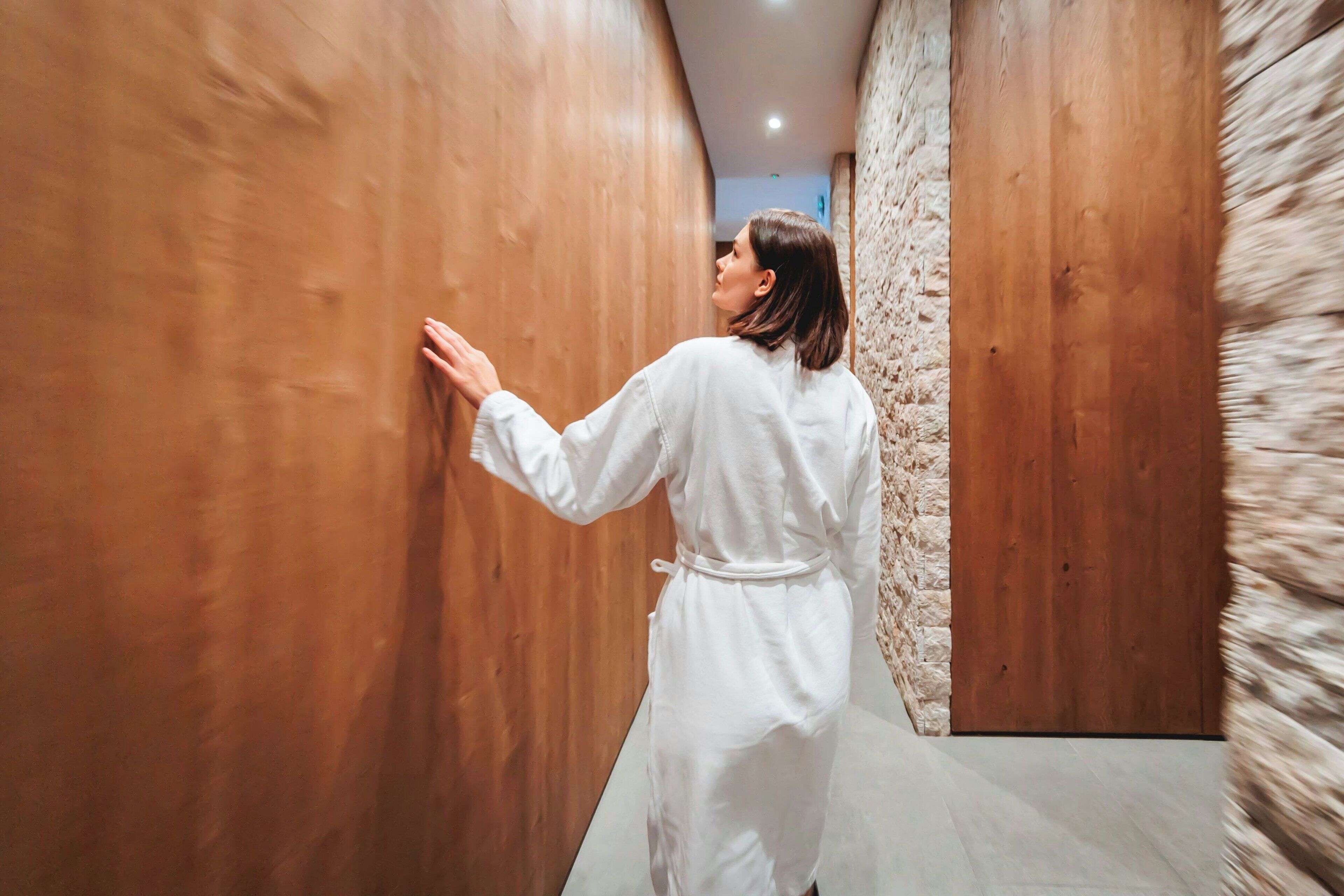 Une personne vêtue d’une longue robe blanche marche dans un couloir aux murs en bois et dont l’un des murs est recouvert de pierres apparentes.