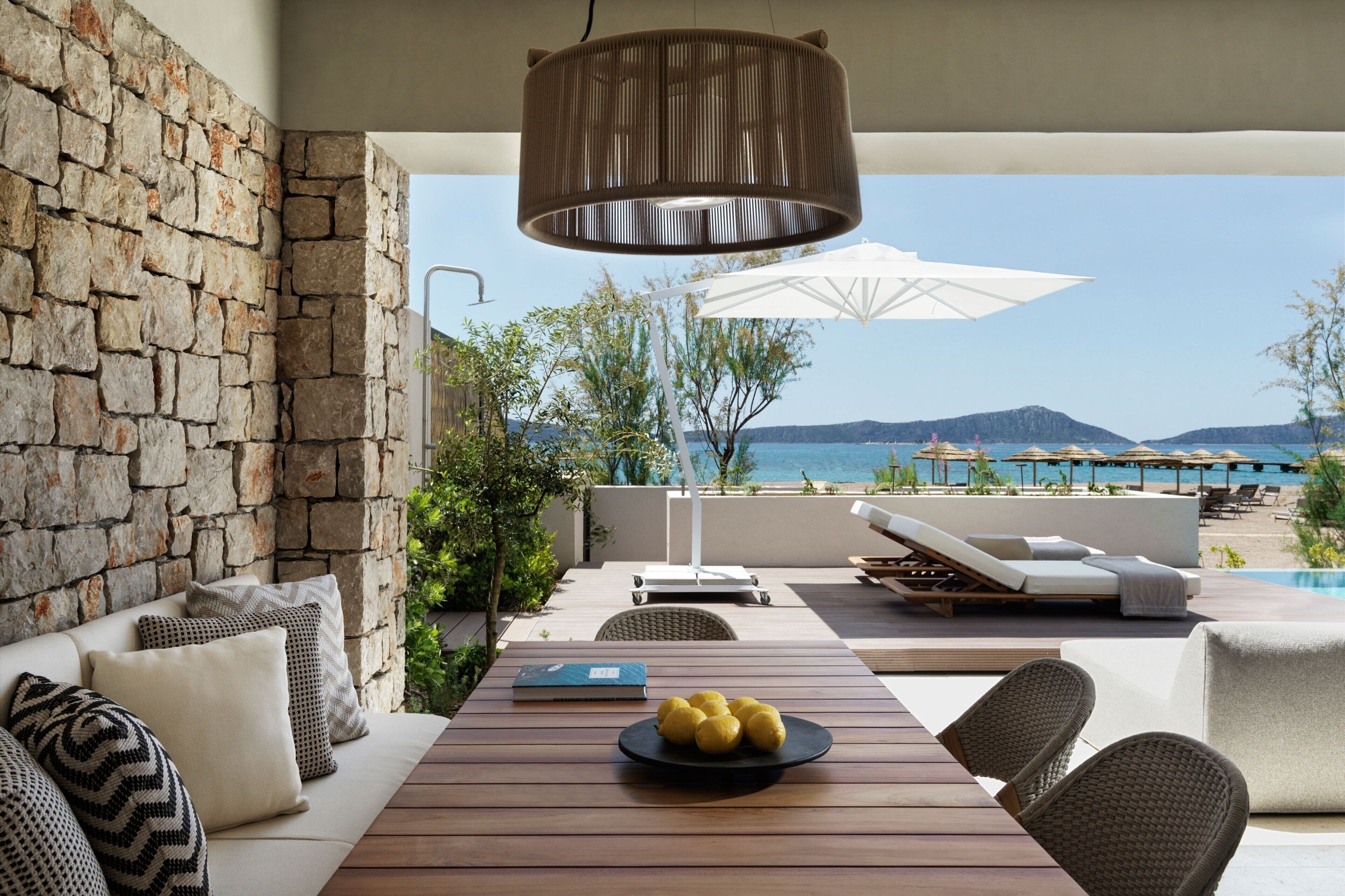A modern outdoor seating area with a stone wall, a wooden table with a plate of lemons, and a view of the sea and mountains.