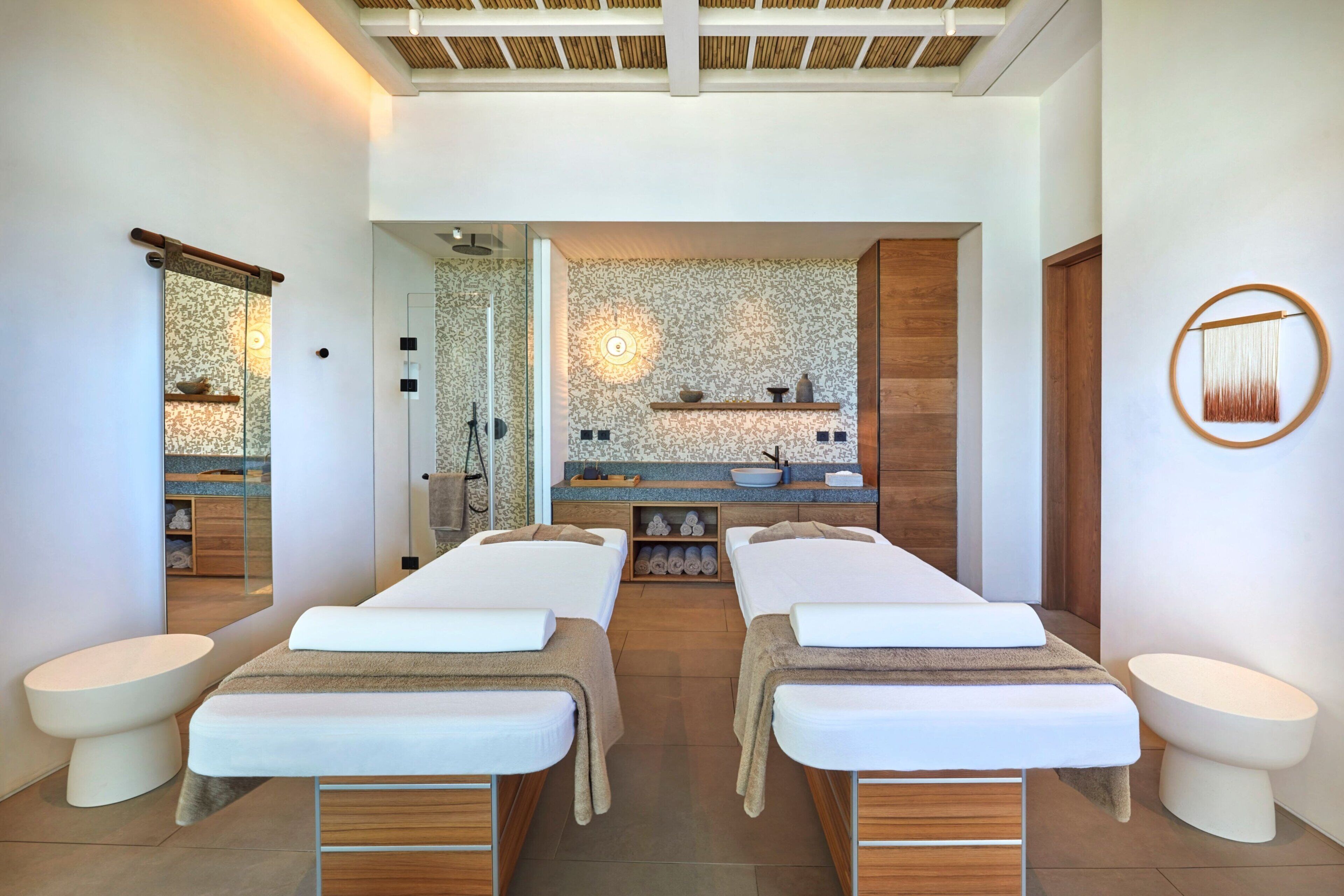 A spa room with two massage tables, a wooden reception desk, a wall-mounted shelf with decorative items, and a large mirror.