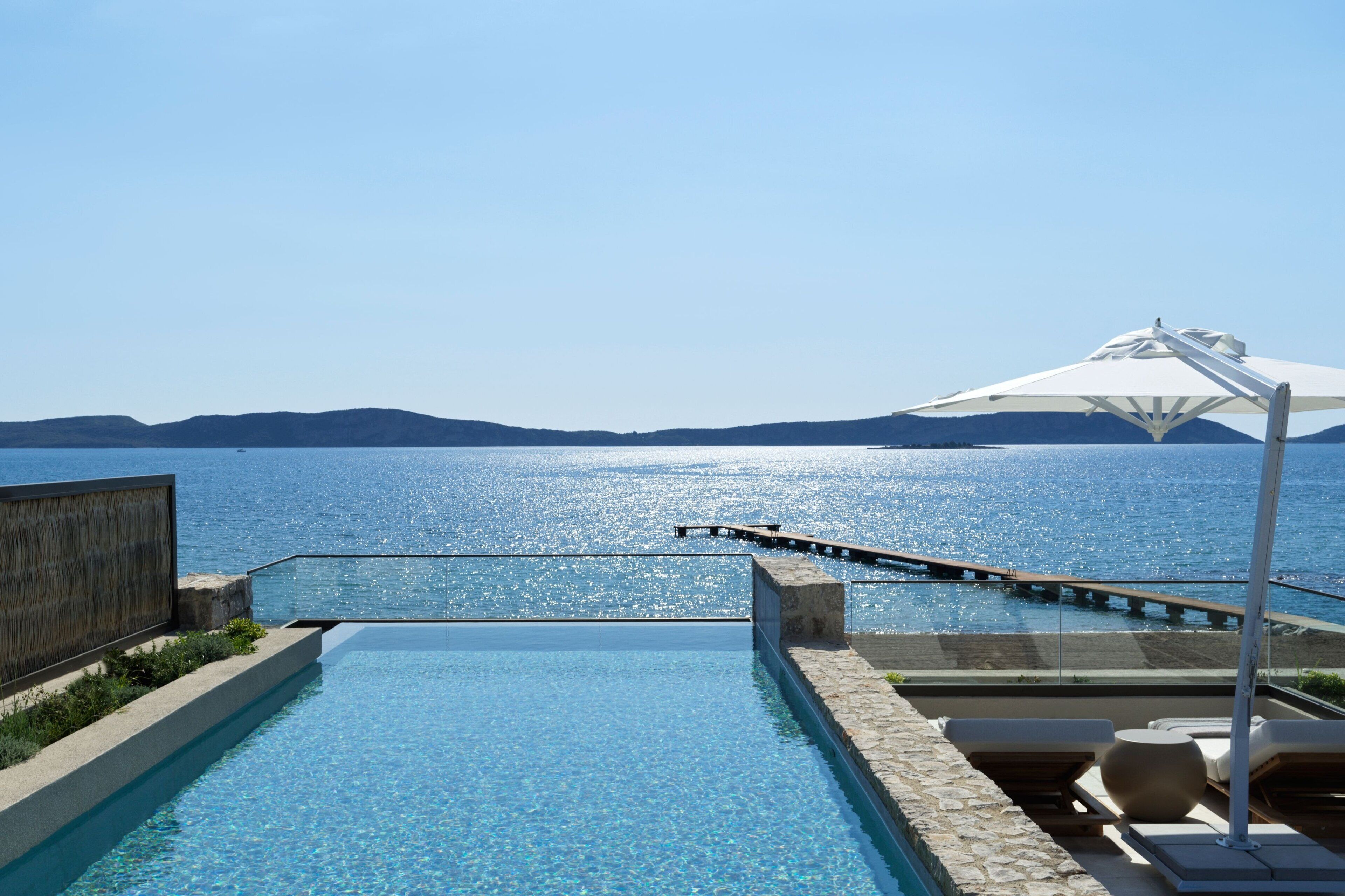 A swimming pool with a view of the sea and mountains, a wooden deck, and lounge chairs.