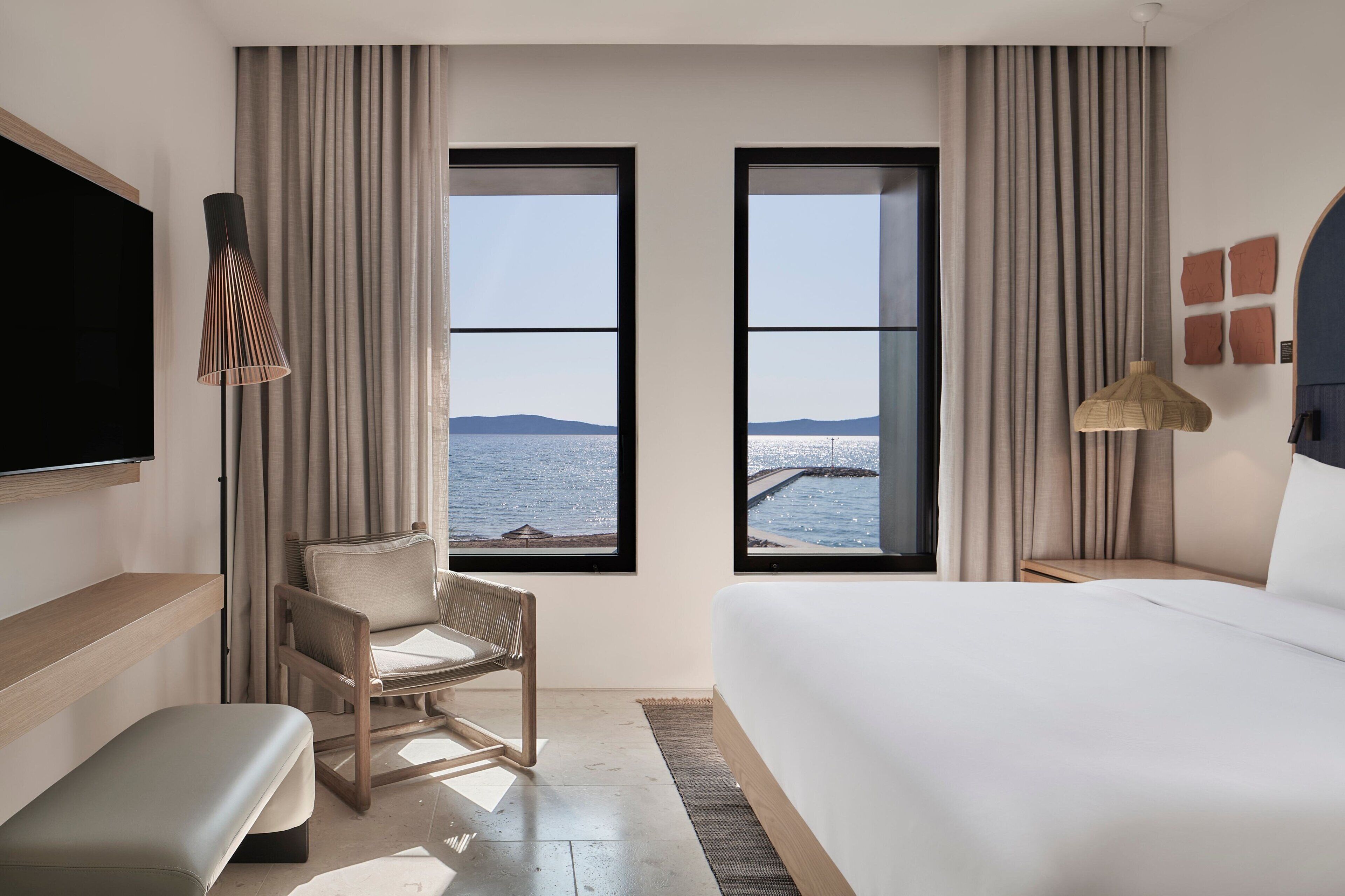 Una habitación de hotel moderna con una cama grande, una silla, un banco, un televisor y dos ventanas con vistas al mar.