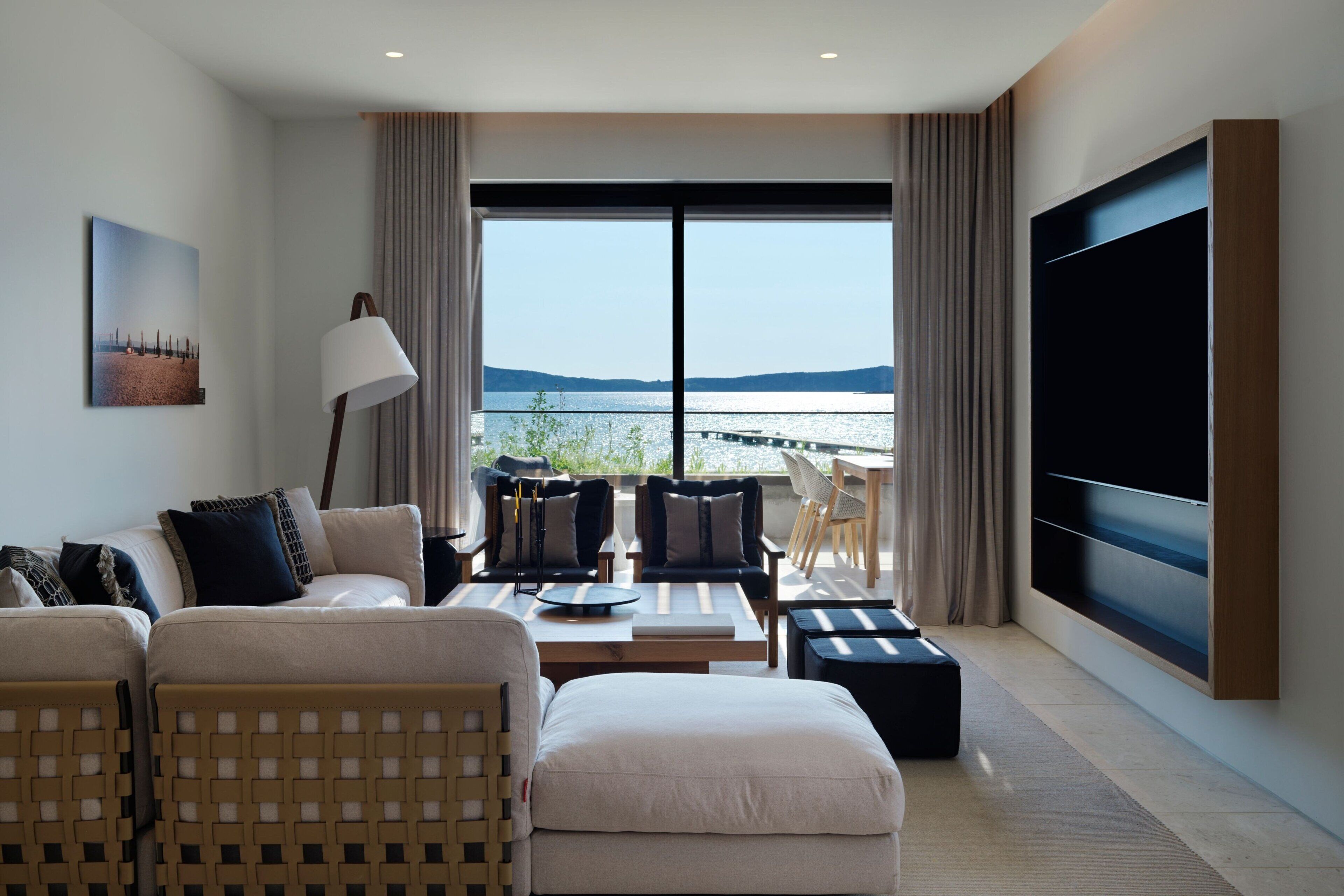 A modern hotel room with a sofa, armchairs, a coffee table, and a TV. The room has a large window with a view of the sea and hills.