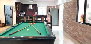 A pool table with a green felt surface and pool cues, a kitchen area with dark wooden cabinets and a microwave, and a brick wall with a window.