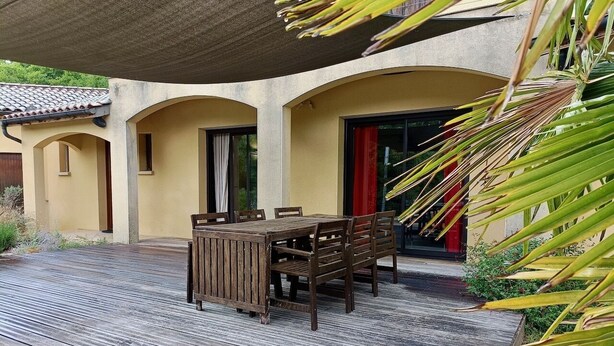 Eine Holzterrasse mit Tisch und Stühlen, ein überdachter Bereich und ein Gebäude mit Rundbögen und Fenstern.
