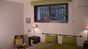 Un dormitorio con cama, una silla, una mesita de noche, una ventana con vista a árboles y un aire acondicionado instalado en la pared.