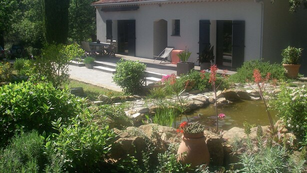 Un edificio blanco con techo de tejas, un patio con mobiliario exterior y un jardín con un estanque.