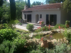 Un edificio bianco con tetto in tegole, un patio con mobili da esterno e un giardino con uno stagno.