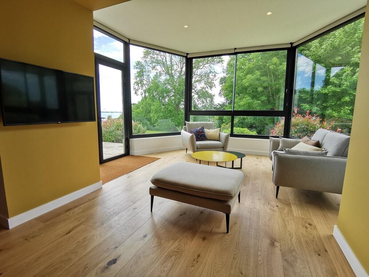 A spacious room with a large window, a flat-screen TV mounted on the wall, a sofa, a coffee table, and an ottoman.