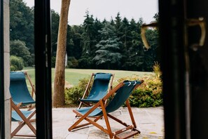 Deux fauteuils de jardin en bois, dotés de coussins bleus, sont placés sur une terrasse en pierre donnant sur un espace verdoyant avec des arbres.