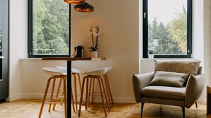 Una cocina moderna con una mesa de comedor blanca, dos taburetes de bar, una lámpara colgante y vistas al exterior a través de amplias ventanas.