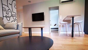 Ein modernes Wohnzimmer mit einer Couch, einem runden schwarzen Tisch, einem an der Wand befestigten Fernseher und einem Essbereich mit Hockern.