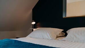 Ein ordentlich bezogenes Bett mit weißen Laken und einer blauen Decke, eine Nachttischlampe und ein dunkles Kopfteil.