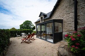 A modern house with a stone exterior, a glass-enclosed patio with outdoor furniture, and a gravel ground. - Le Sant C'hireg    . (Concarneau)