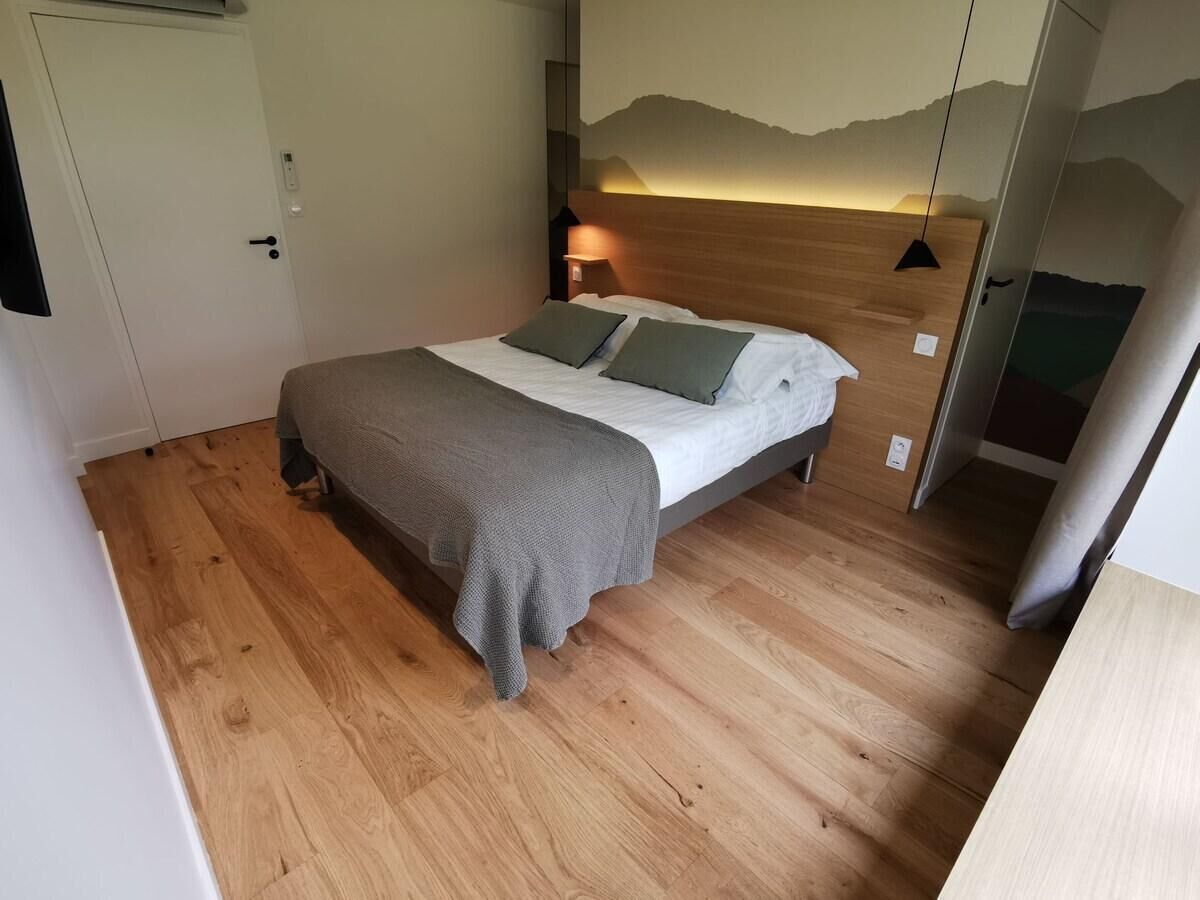A bedroom with a bed, wooden flooring, a wall-mounted headboard, and a nightstand with a lamp.