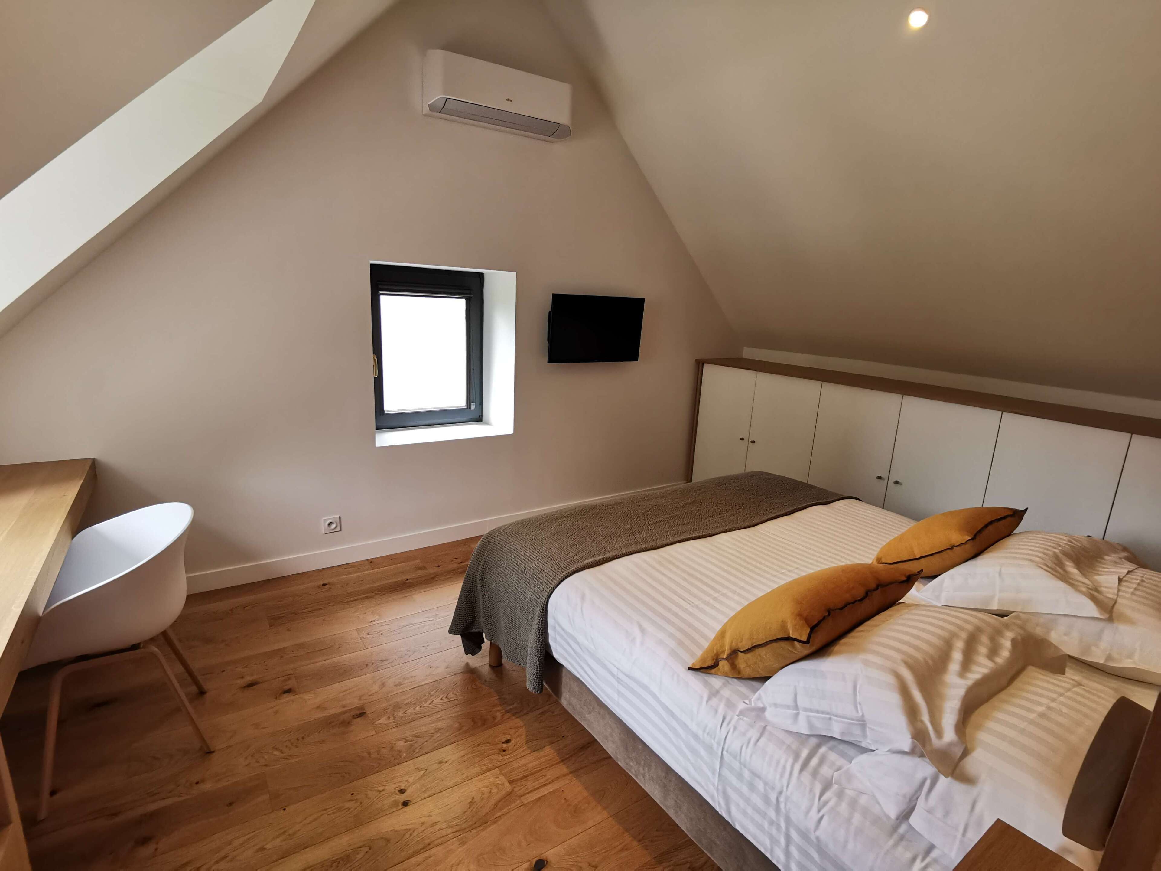 A bedroom with a bed, a desk, a window, and an air conditioning unit.