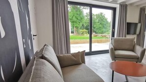 Ein modernes Wohnzimmer mit einer Couch und einem Sessel, einem kleinen Tisch und einer großen Schiebeglastür, die zu einem Garten führt.