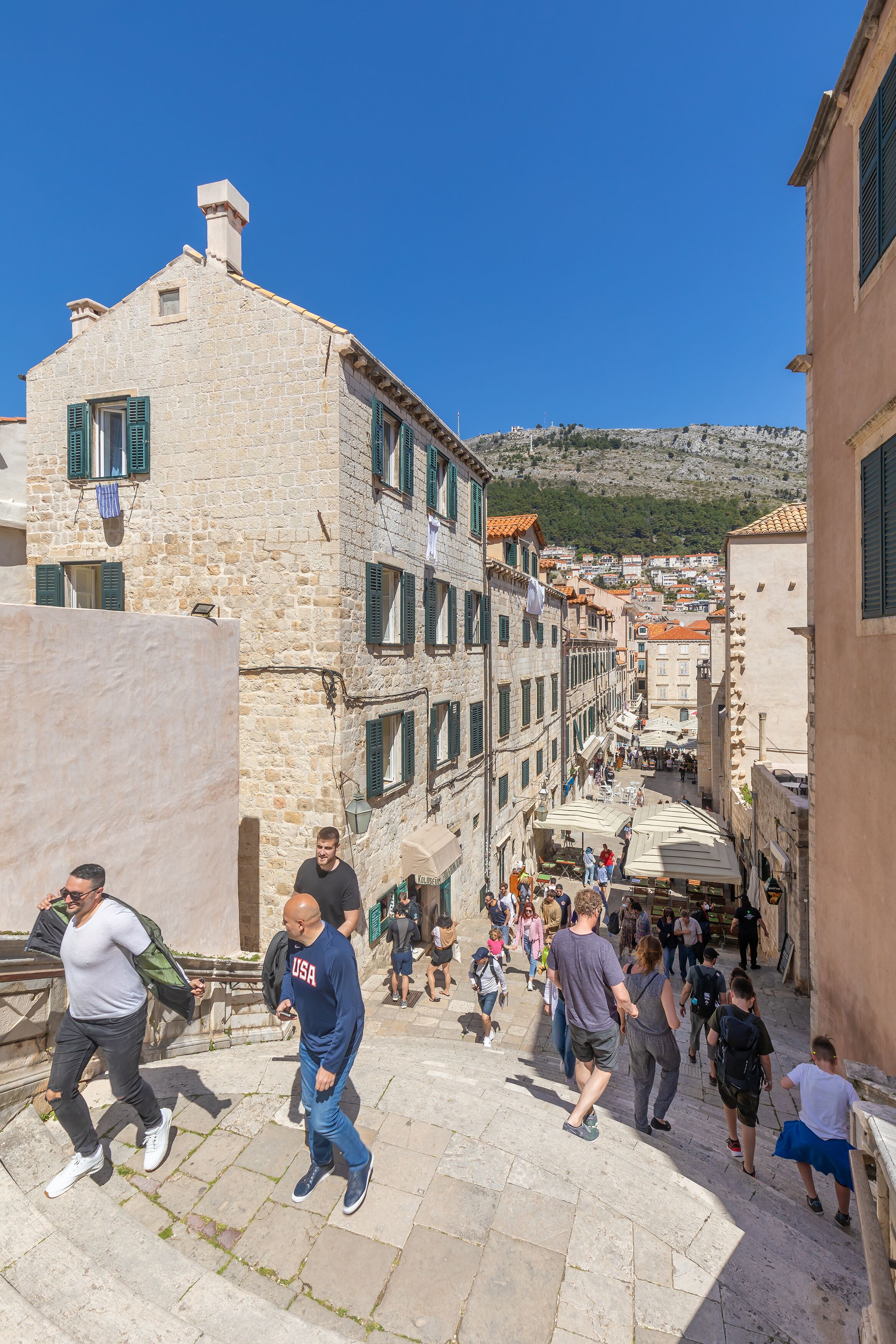 A narrow street with stone buildings, outdoor seating, and people walking.