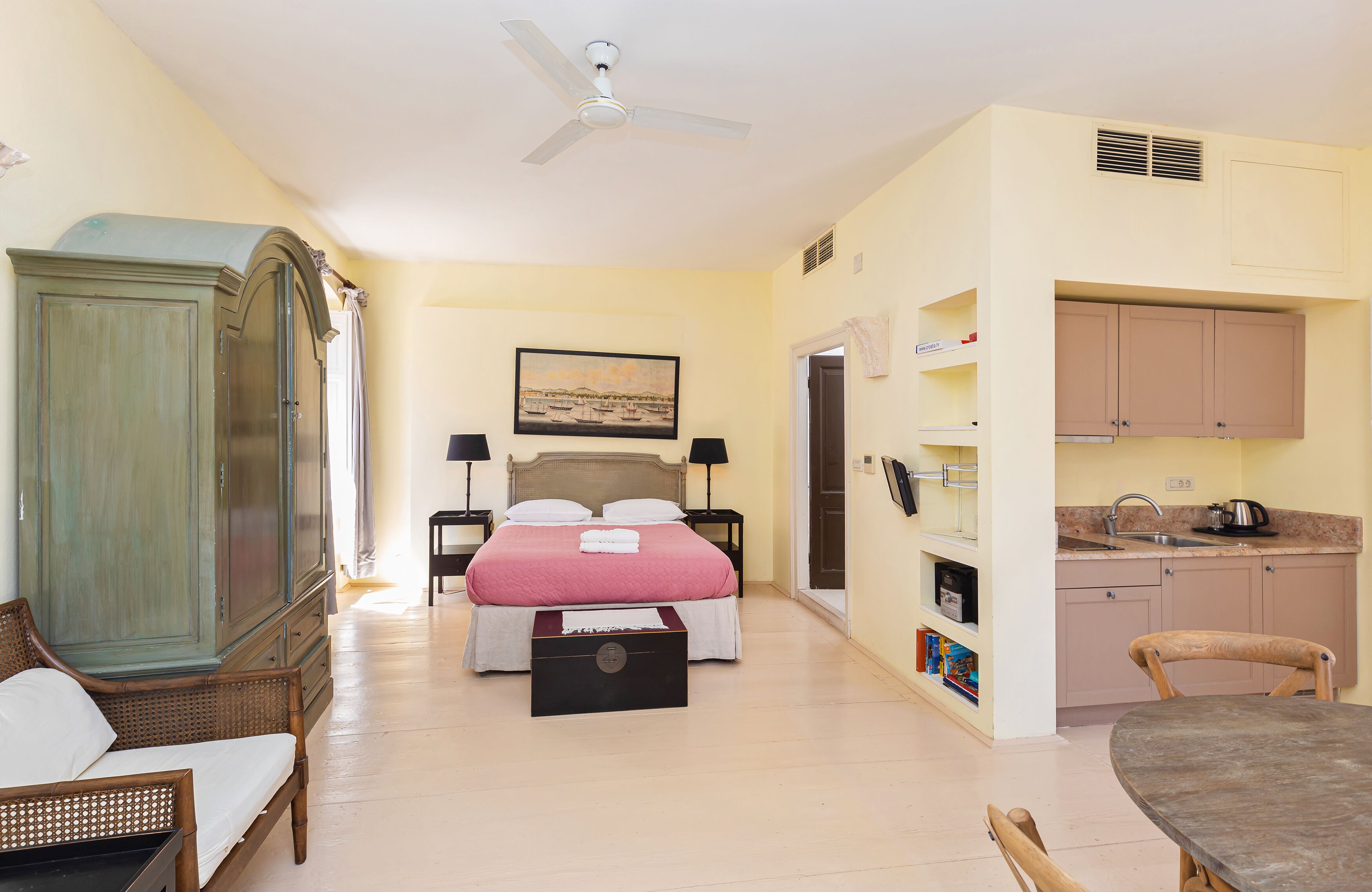 A bedroom with a bed, a wardrobe, a desk, and a kitchenette.