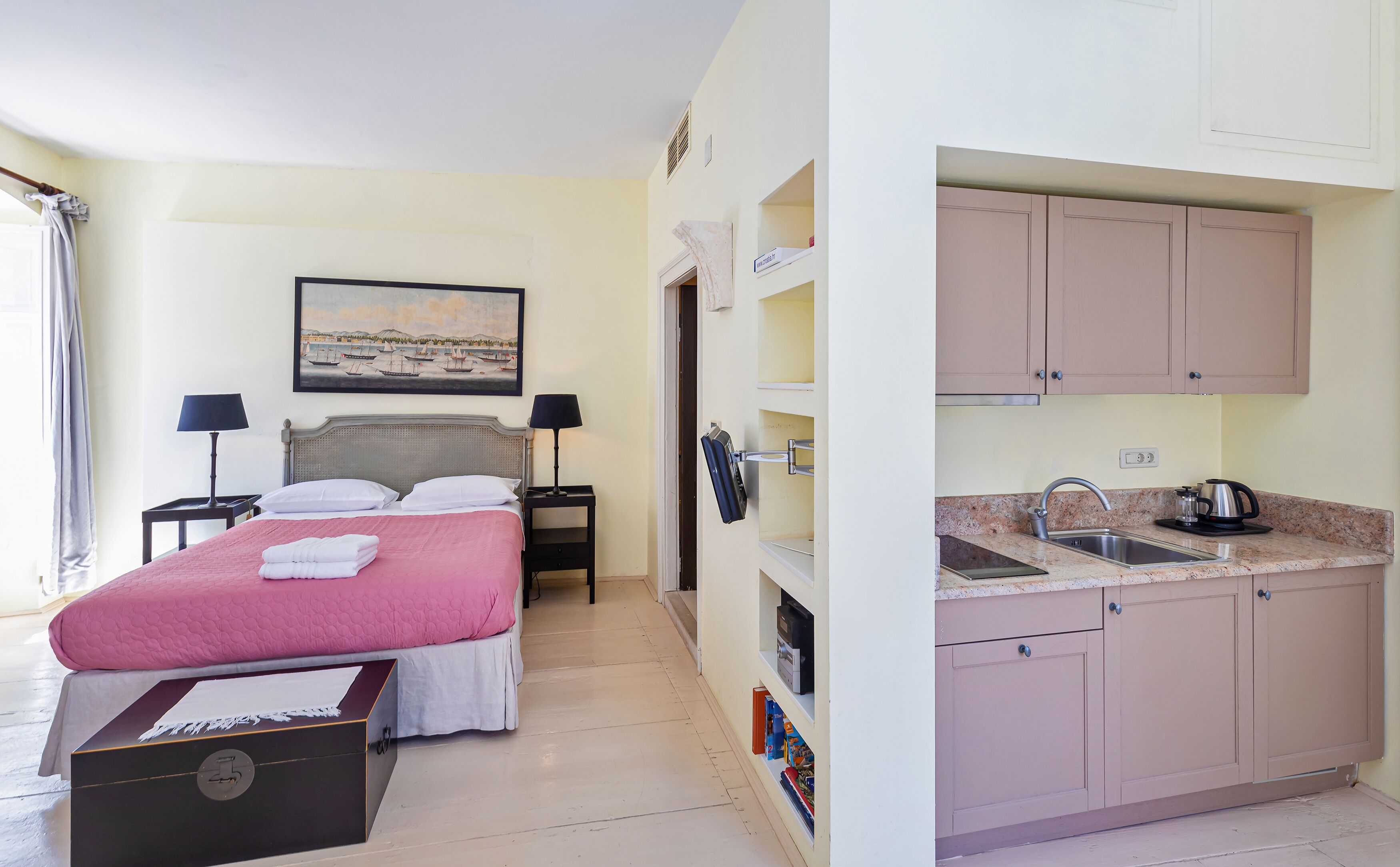A hotel room with a bed, bedside tables, a painting, a wardrobe, and a kitchenette.
