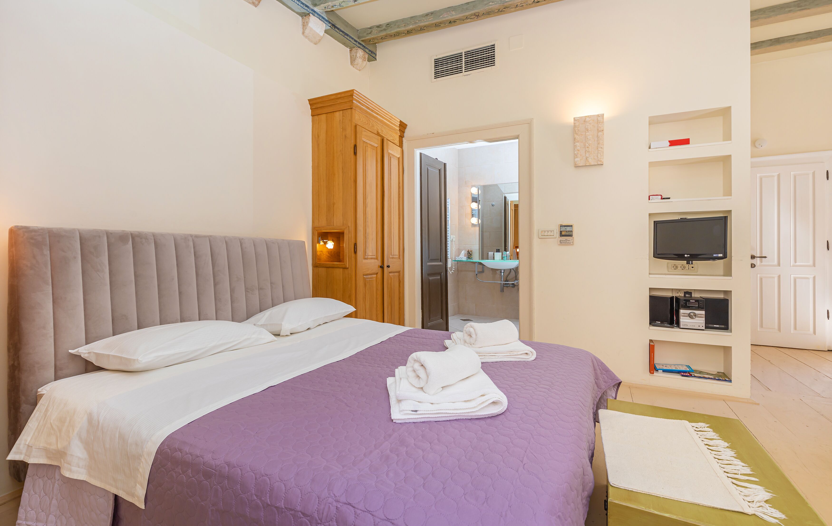 A bedroom with a large bed, a wooden wardrobe, a television, and a bathroom visible through an open door.