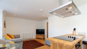 A modern living room with a sofa, a glass coffee table, a TV, and a kitchen area with a wooden countertop and a sink.