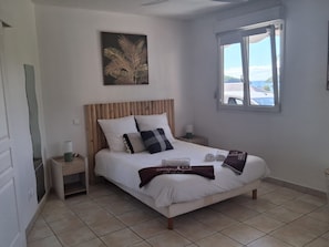 A bedroom with a bed, a nightstand, a window, and a wall art piece. - T2 Anse à l'Ane, a 5-minute walk from the beach (Les Trois-Îlets)