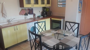 Una cocina compacta con armarios amarillos, lavadora, microondas y una zona de comedor con mesa y sillas de vidrio.