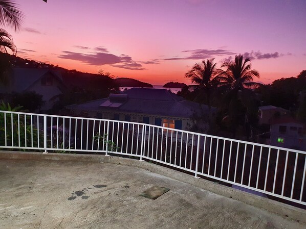 A balcony with a concrete floor and white railing overlooking a residential area with houses and palm trees during sunset. - T2 Anse à l'Ane 5-minute walk from the beach (Les Trois-Îlets)