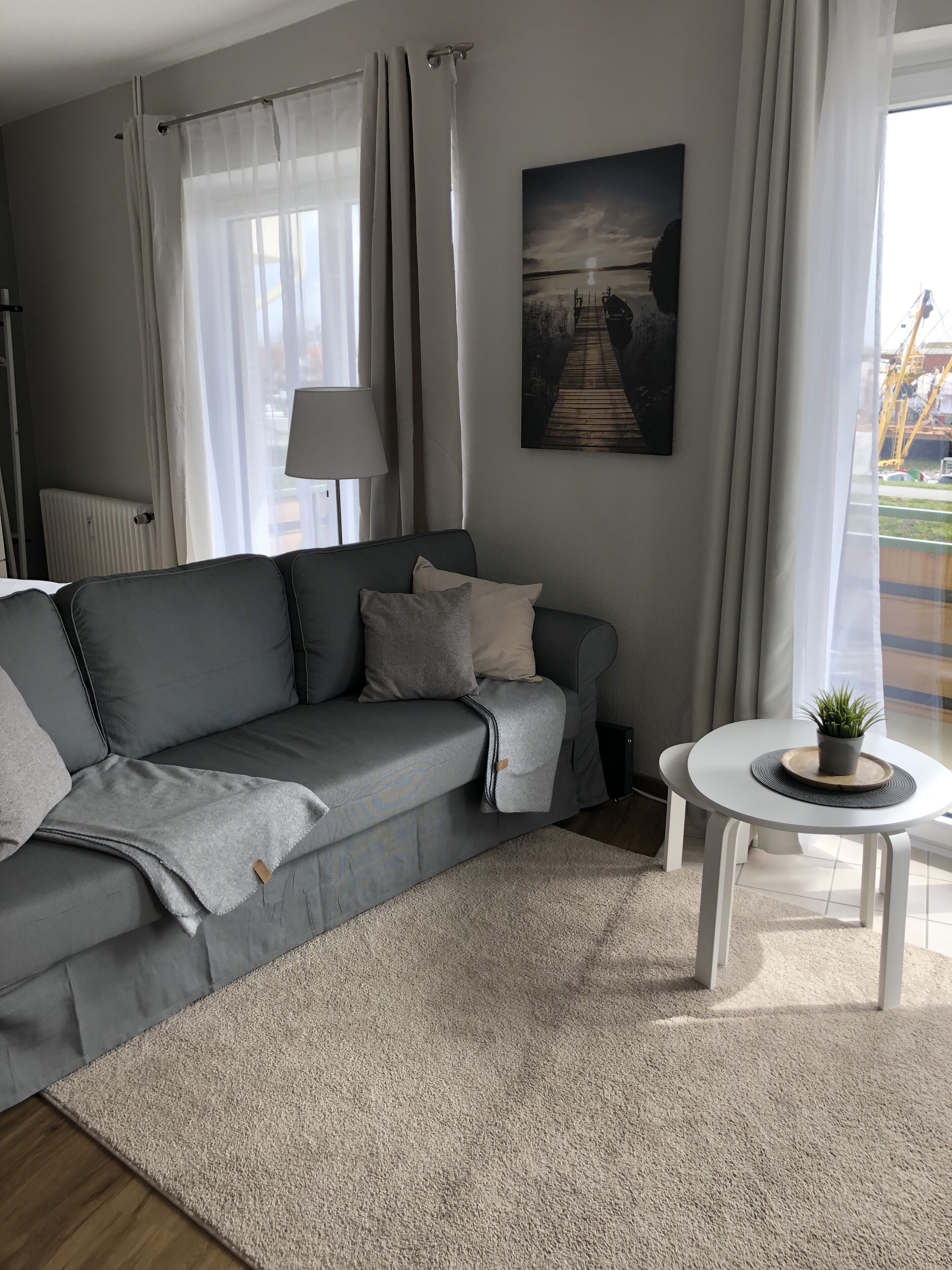 A modern living room with a grey sofa, a small white table with a plant, a beige rug, and a painting of a bridge on the wall.