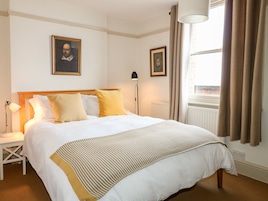 Una cama bien tendida con ropa de cama blanca y mostaza, una lámpara de noche y dos cuadros en la pared.