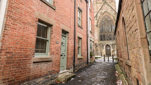 Une ruelle étroite bordée d’immeubles en briques rouges, une église surmontée d’un clocher en flèche et une entrée fermée par un portail.