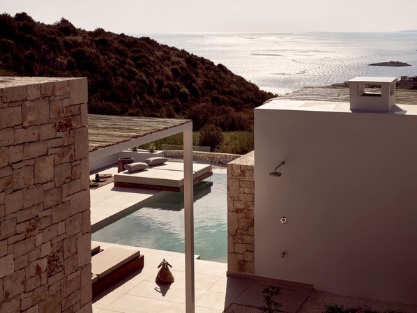 Un espace piscine extérieur moderne, agrémenté d’un mur en pierre, d’un coin salon avec des coussins et offrant une vue sur la mer.