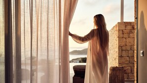 A person in a white dress standing by a window with sheer curtains, overlooking a stone wall and a view of the sea. - The Sall Suites Complex A - Sea Views (Zakynthos)