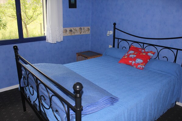 A bedroom with a blue bedspread, a red pillow, a small wooden nightstand, and a window with a view of greenery.