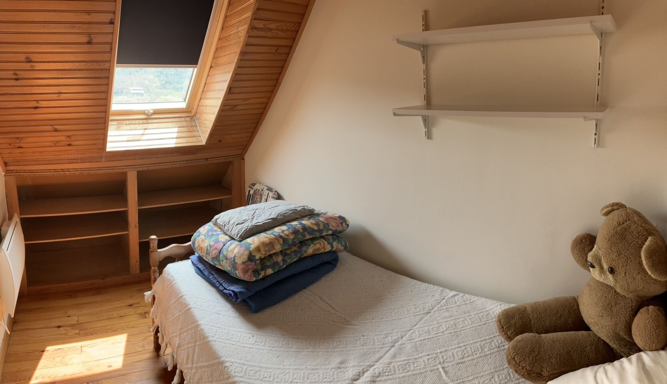 Une pièce chaleureuse avec un plafond en pente, des murs en bois, un lit avec une literie à motifs floraux, un ours en peluche et une étagère blanche.
