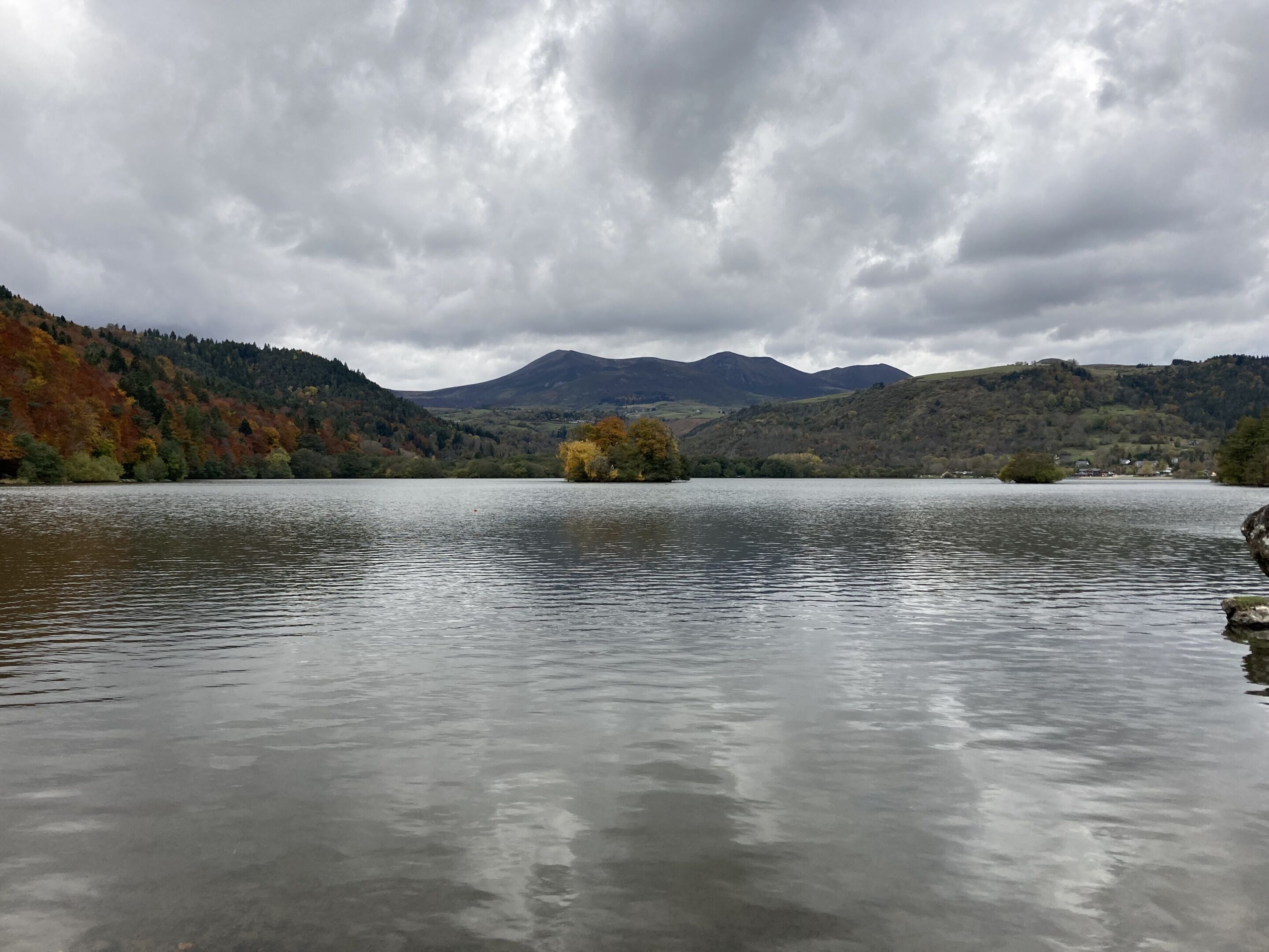 Un lago tranquilo con una cadena montañosa al fondo y árboles de colores otoñales en las colinas.
