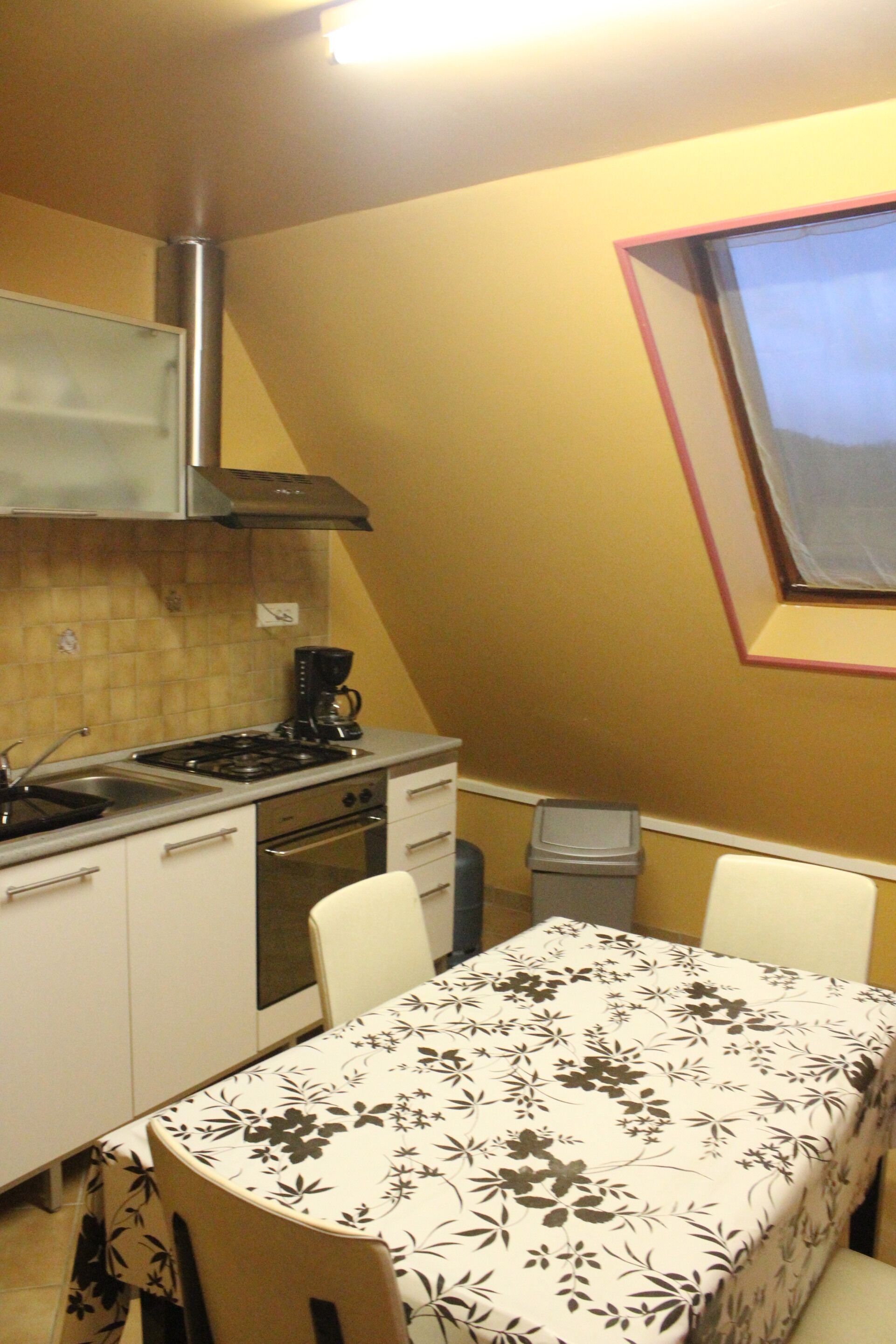 A kitchen with a stove, oven, and sink, a dining table with a floral tablecloth, and a window with a pink frame.