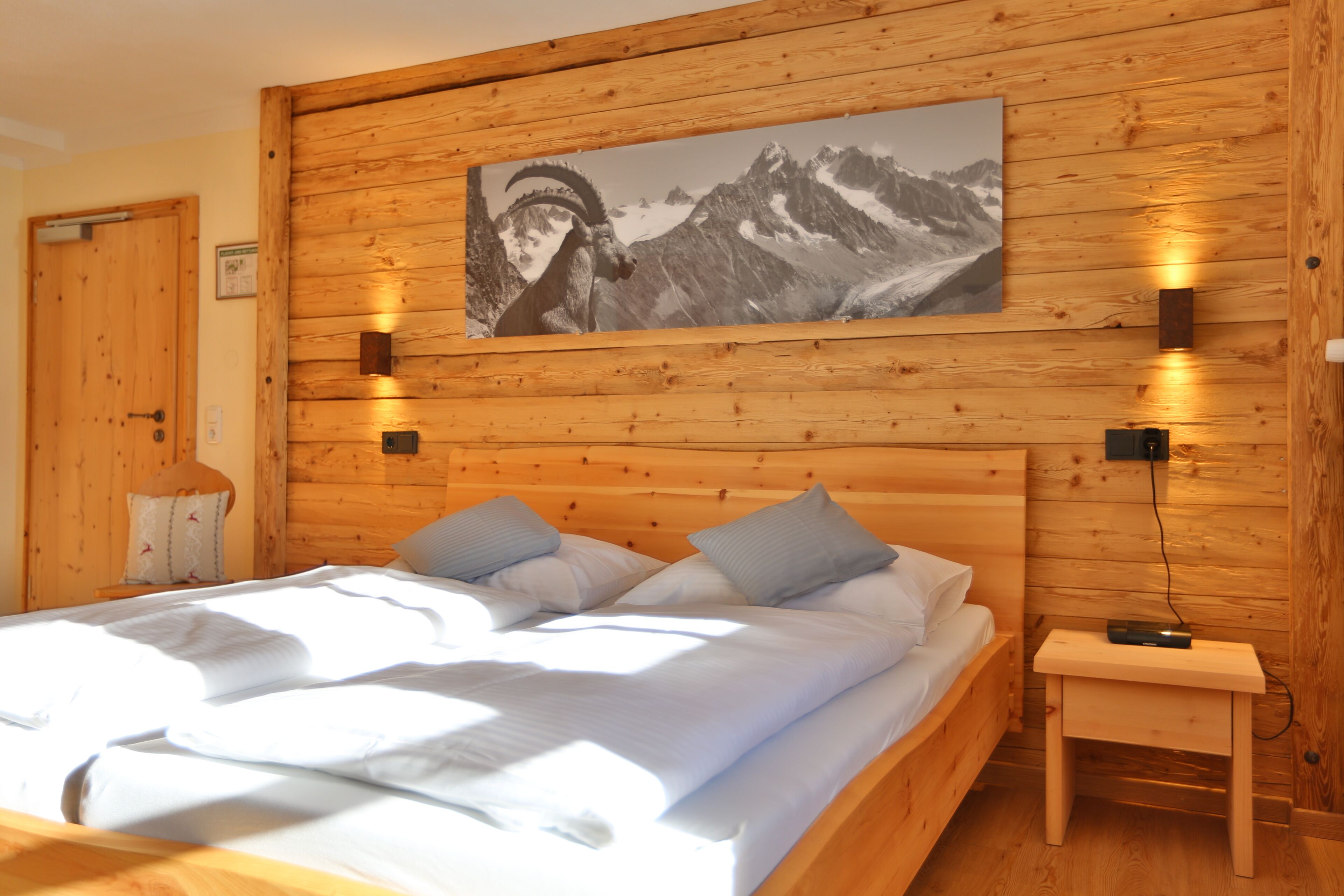 A wooden-paneled room with a bed, pillows, a nightstand, a wall-mounted picture, and a door.