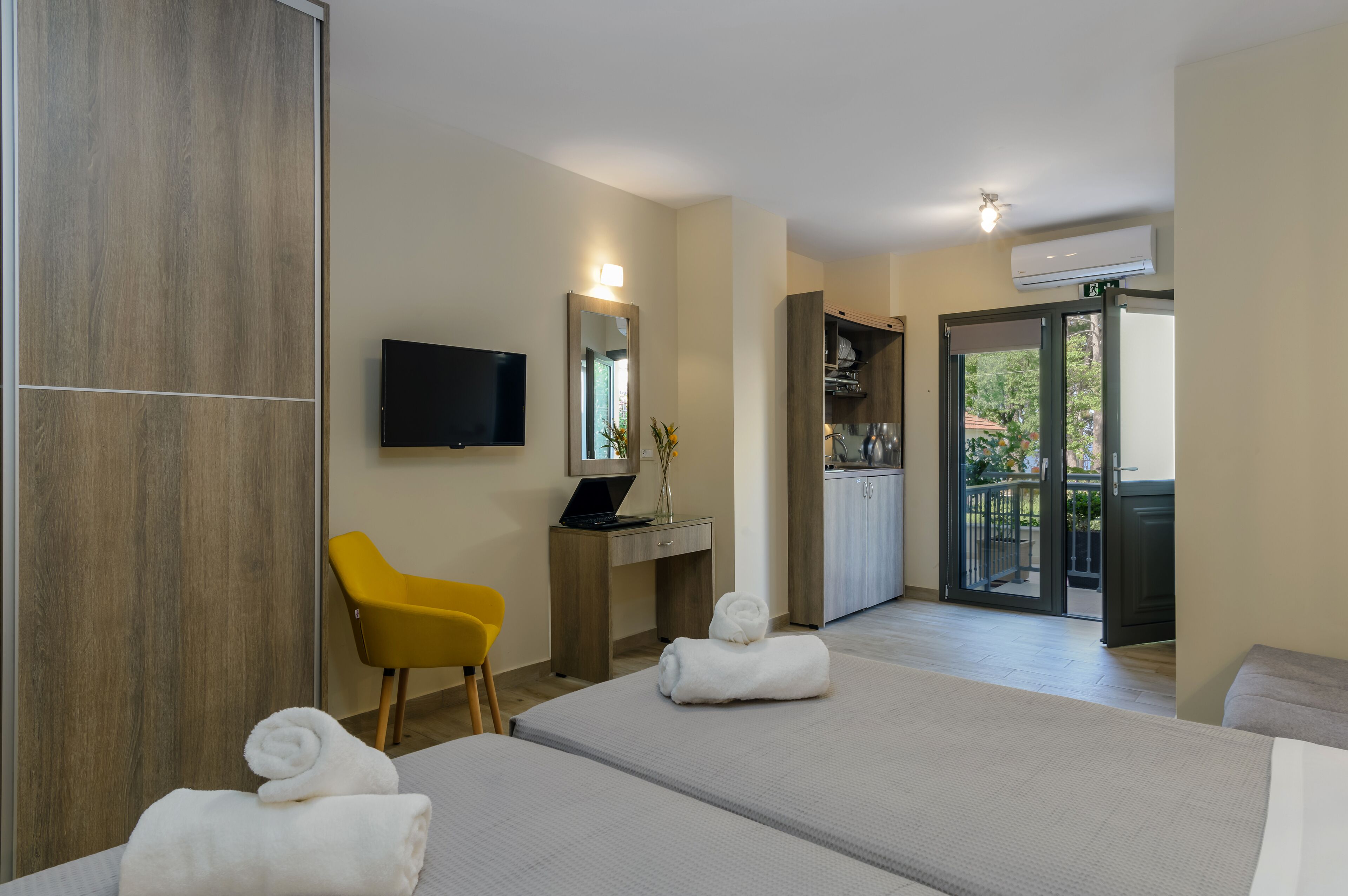 Ein Hotelzimmer mit zwei Betten, einem Schreibtisch mit Computer, einem gelben Stuhl, einem Fernseher und Blick auf die Küche und den Balkon.