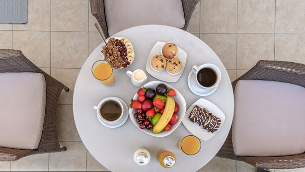 Ein runder Tisch mit Frühstücksleckereien wie Früchten, Muffins und Kaffee, umgeben von Korbstühlen.
