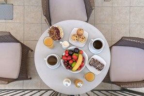 Mesa redonda com itens de café da manhã, incluindo frutas, muffins e café, cercada por cadeiras de vime.